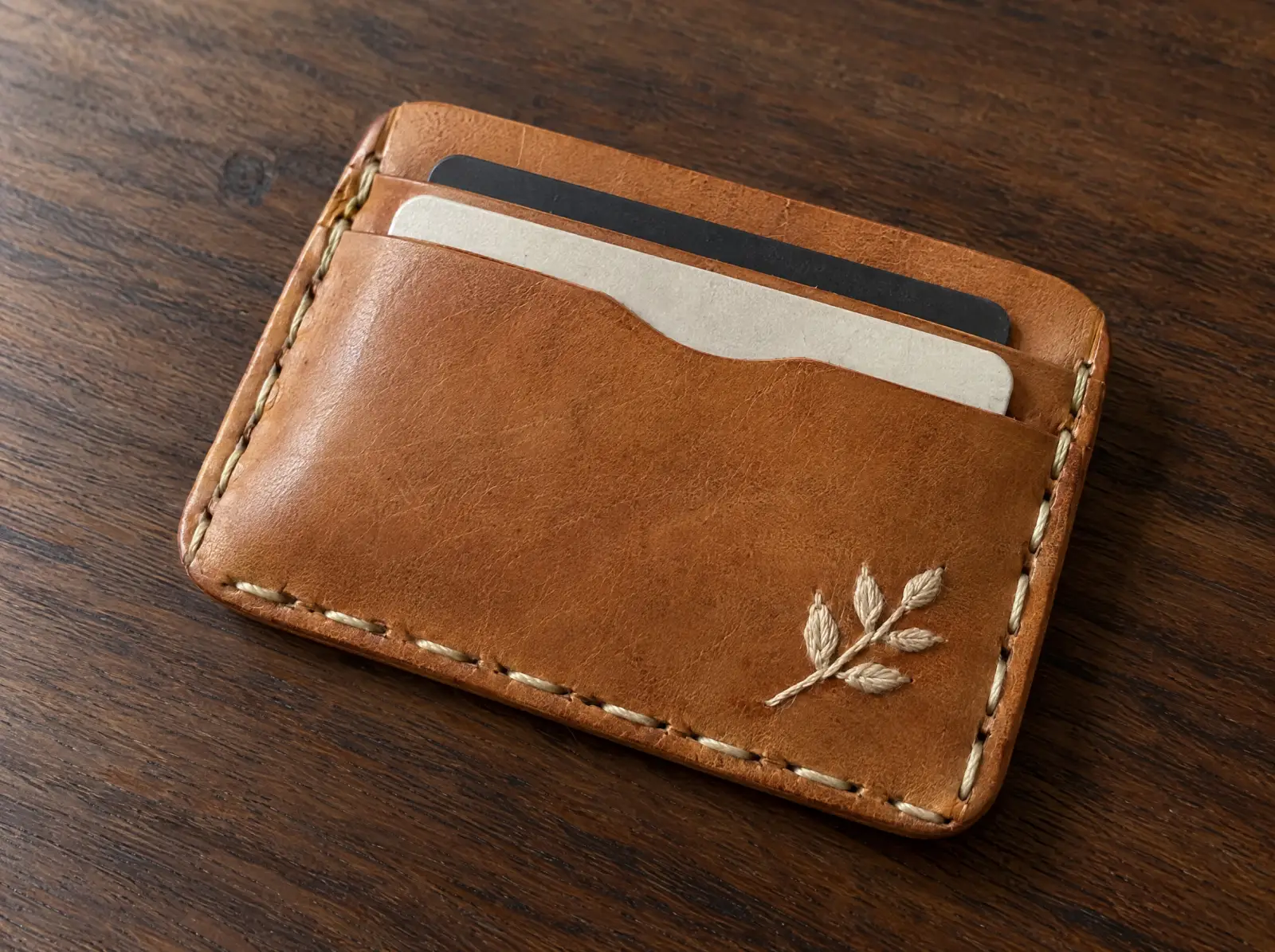 A tan leather card wallet with a small cream embroidered leaf motif